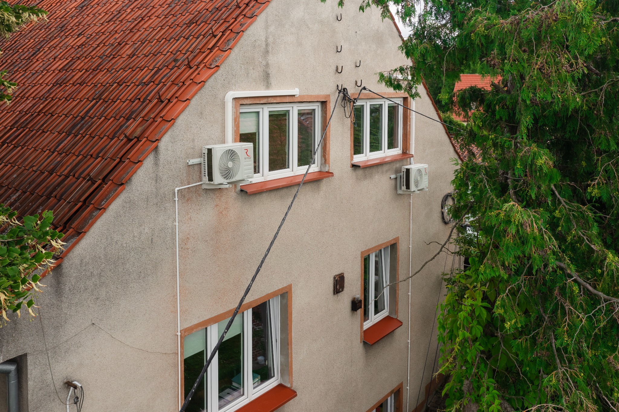 Dwa klimatyzatory zewnętrzne zamontowane na elewacji dwupiętrowego domu z czerwoną dachówką, widoczne okna z brązowymi parapetami, oplecione kable elektryczne i fragmenty zieleni drzew.