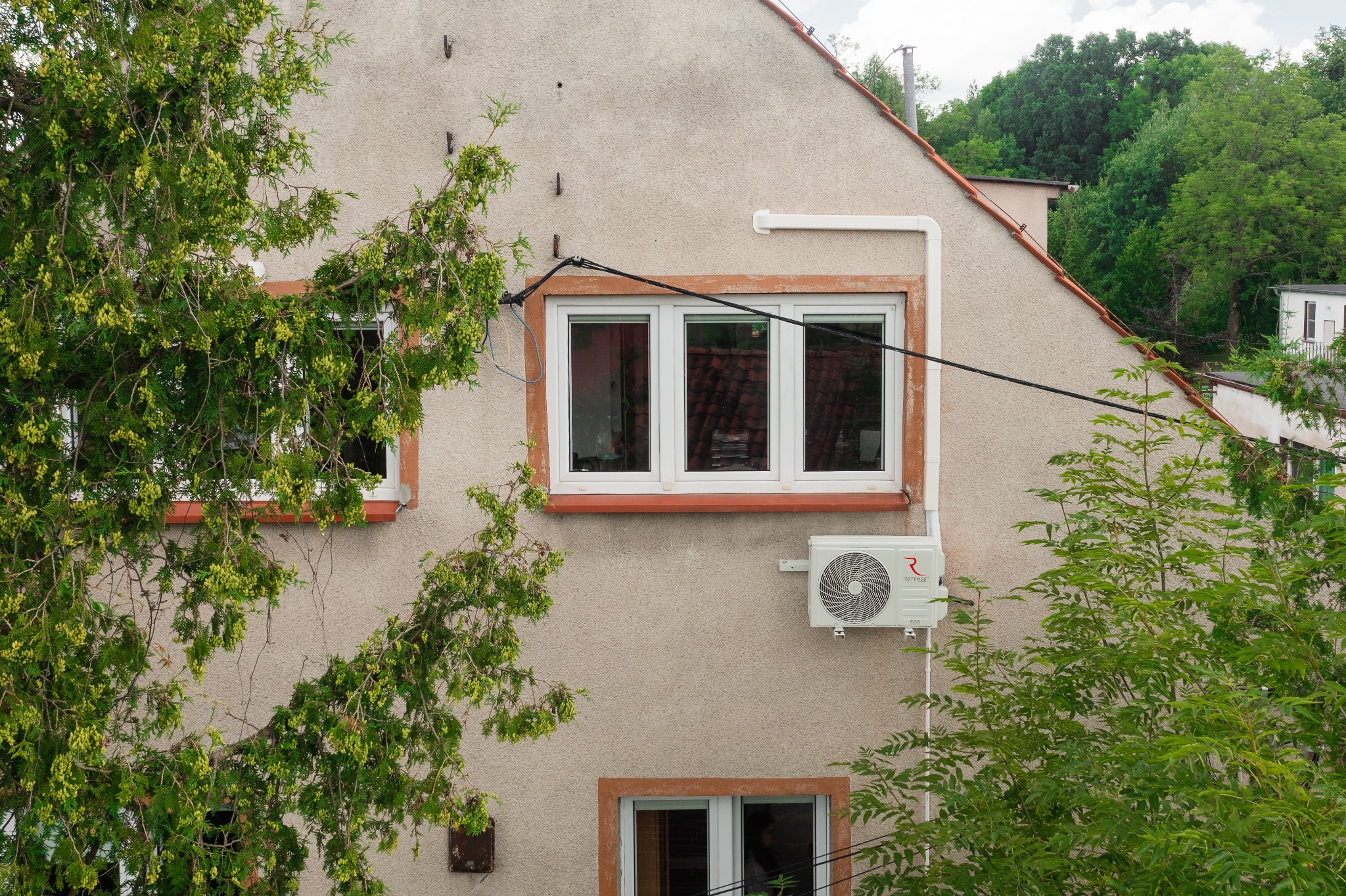 Zewnętrzna jednostka klimatyzacji Rotenso zamontowana na elewacji domu, widoczna spod gałęzi drzewa, z oknem w tle.