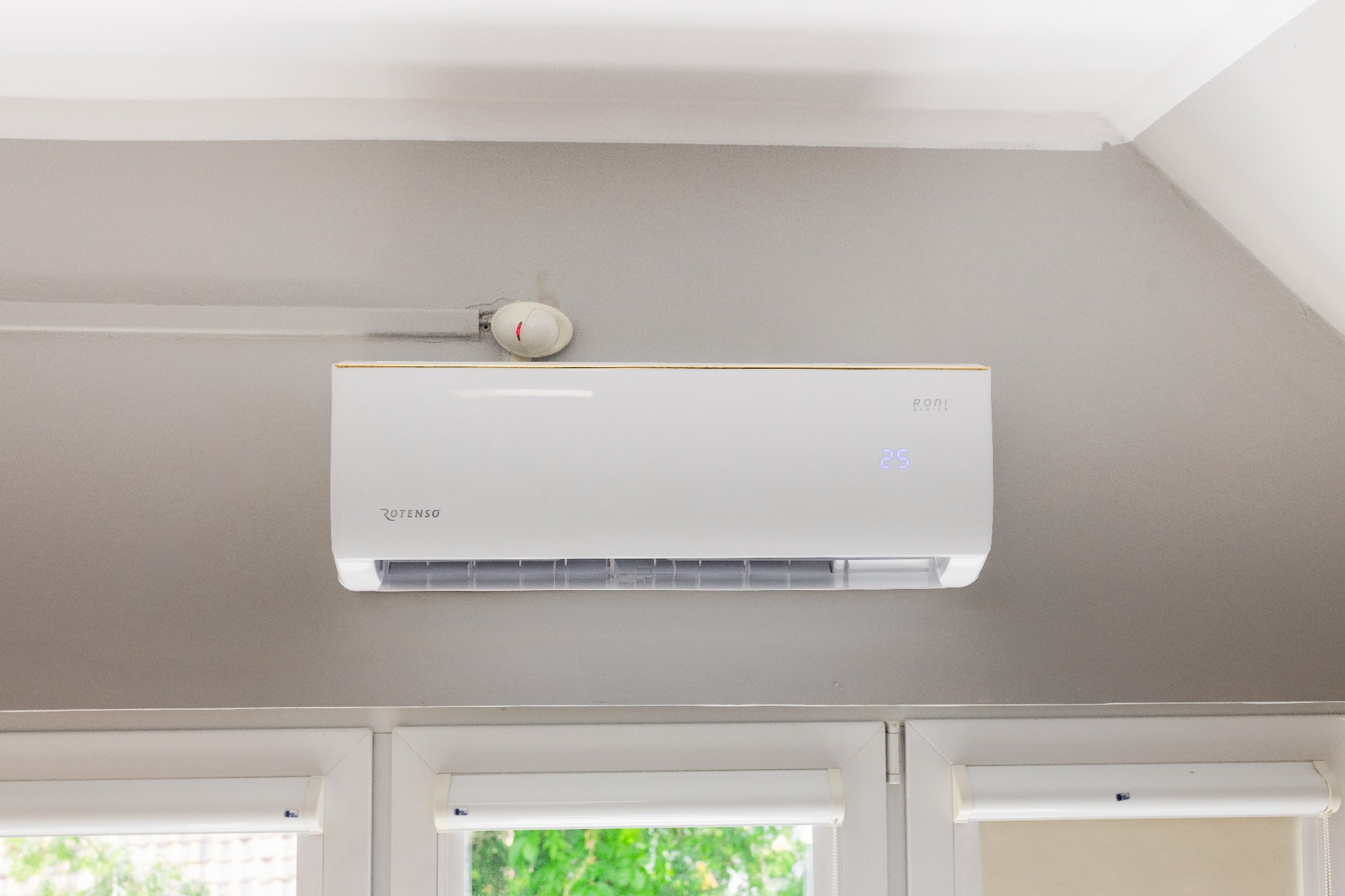 Biała klimatyzacja Rotenso zamontowana na szarej ścianie nad oknem z roletami, widoczny czujnik dymu na suficie.
