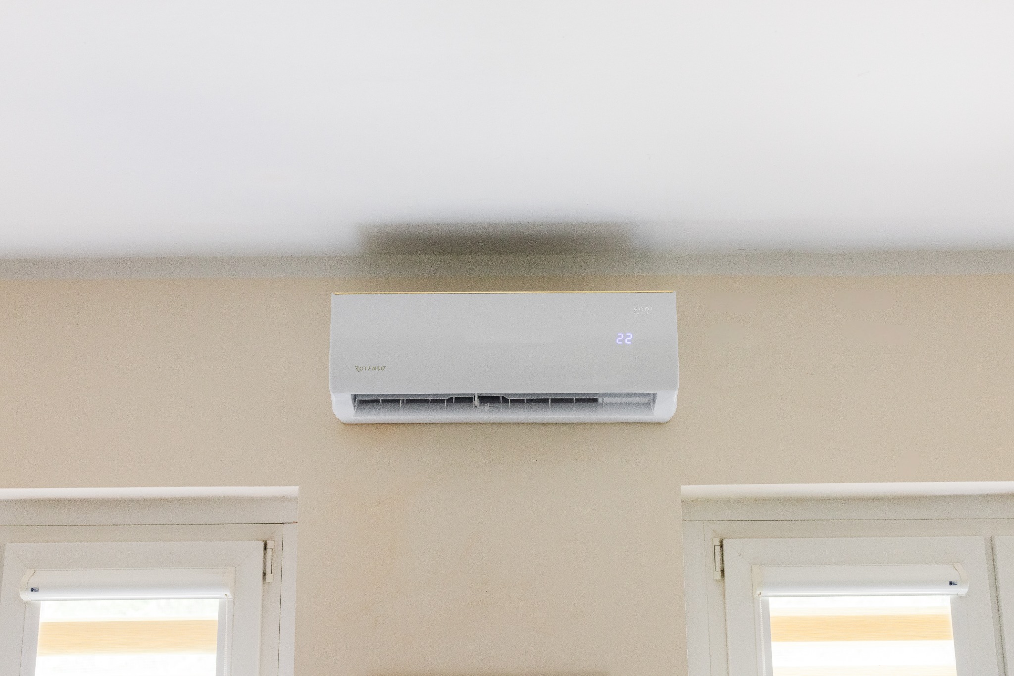 Biały klimatyzator marki Rotenso zamontowany na beżowej ścianie nad dwoma oknami z roletami w pomieszczeniu.