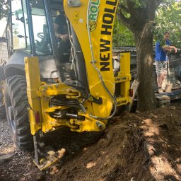 BEL-POL Kamil Bełczącki - Żółta koparko-ładowarka New Holland w trakcie prac ziemnych, widoczny operator w kabinie oraz mężczyzna z łopatą na tle drzewa i konstrukcji budowlanej.