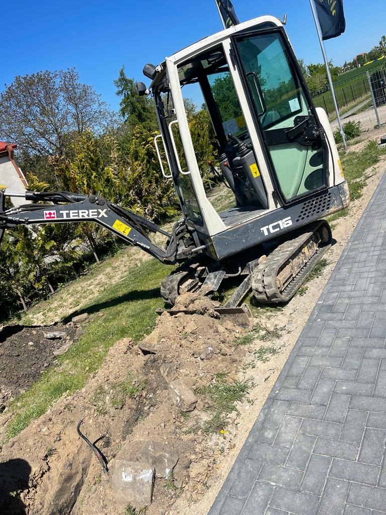 Mała koparka Terex TC16 na gąsienicach podczas prac ziemnych przy chodniku z kostki brukowej, widoczny wykop z kablem w ziemi.