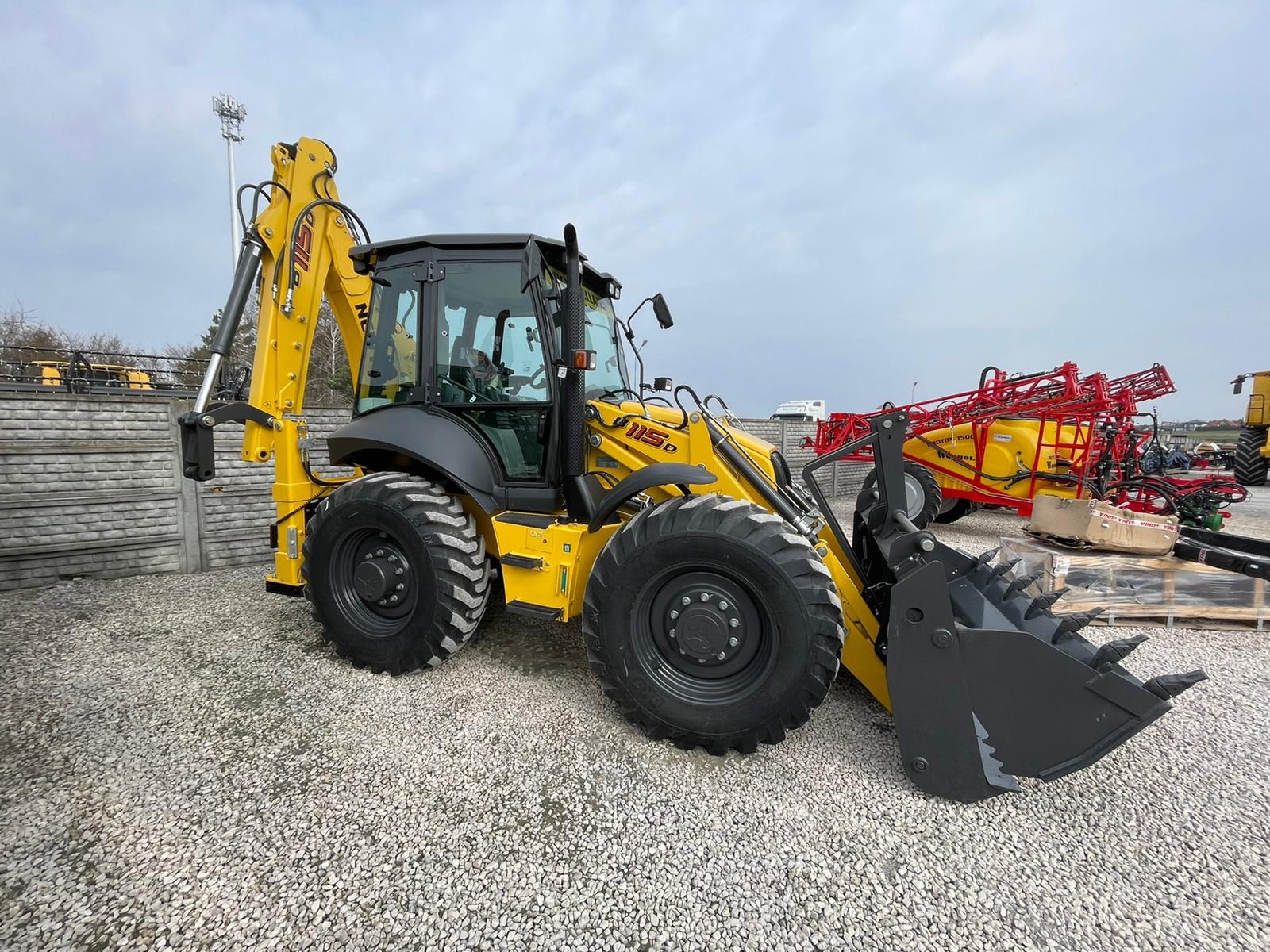 Żółta maszyna budowlana z łyżką z zębami na żwirowym terenie, model 115D, w tle widoczny opryskiwacz rolniczy i betonowe ogrodzenie.