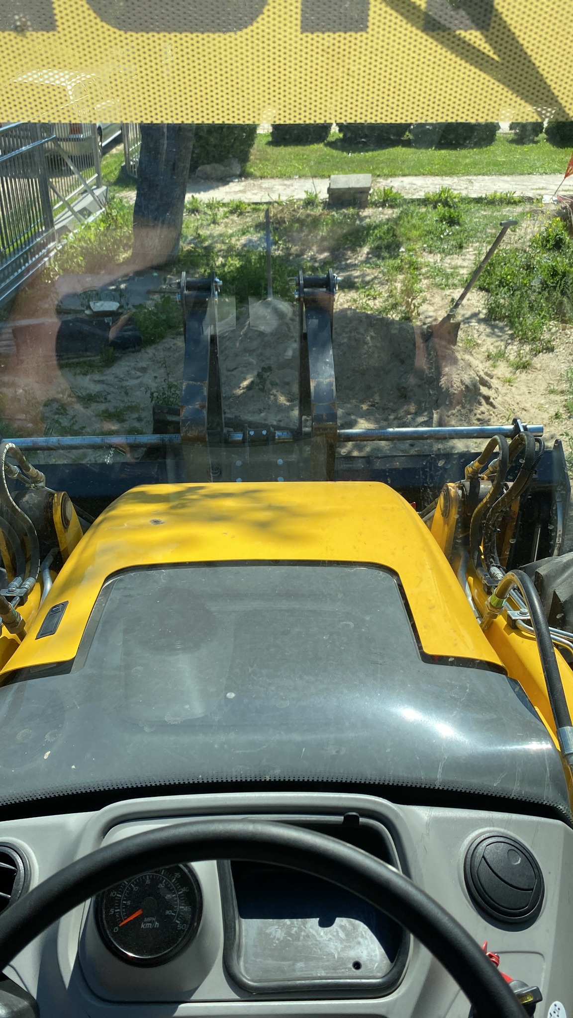 Widok z kabiny żółtej maszyny budowlanej na wykop ziemny i łopatę, widoczne ramię koparki i fragment terenu zielonego.