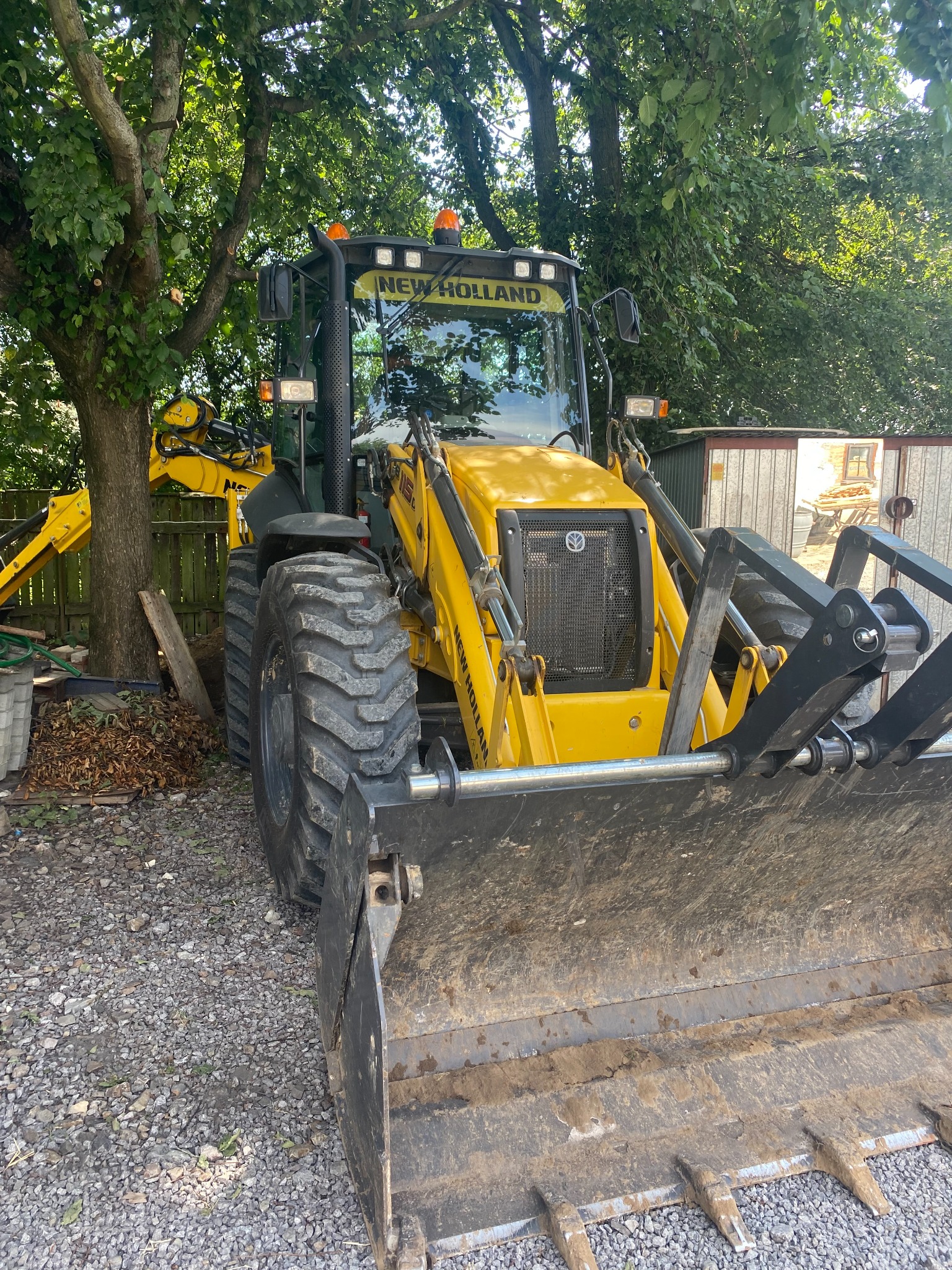 Żółta maszyna budowlana marki New Holland z łyżką, zaparkowana na żwirowym podłożu w cieniu drzew.
