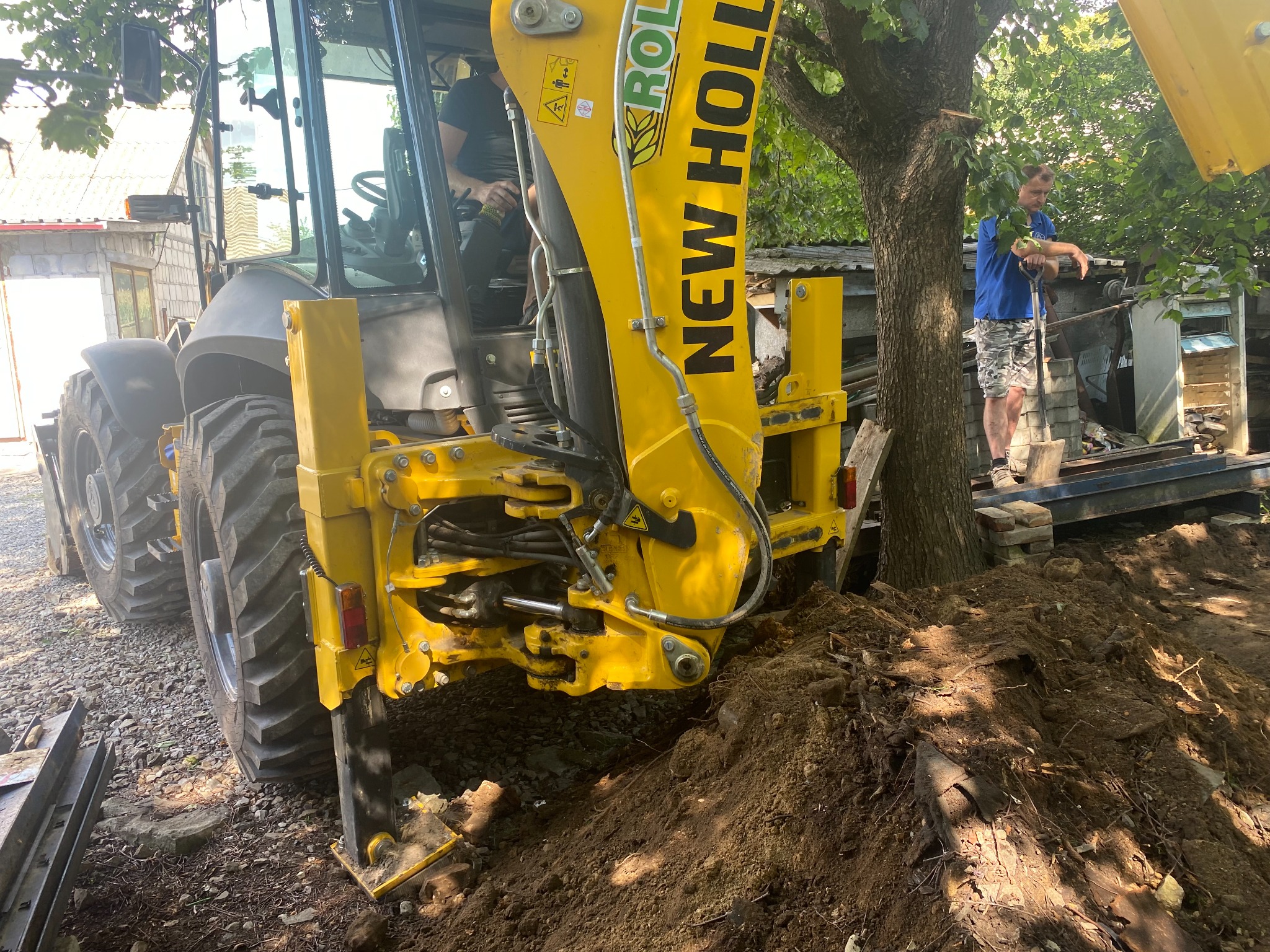 Żółta koparko-ładowarka New Holland w trakcie prac ziemnych, widoczny operator w kabinie oraz mężczyzna z łopatą na tle drzewa i konstrukcji budowlanej.