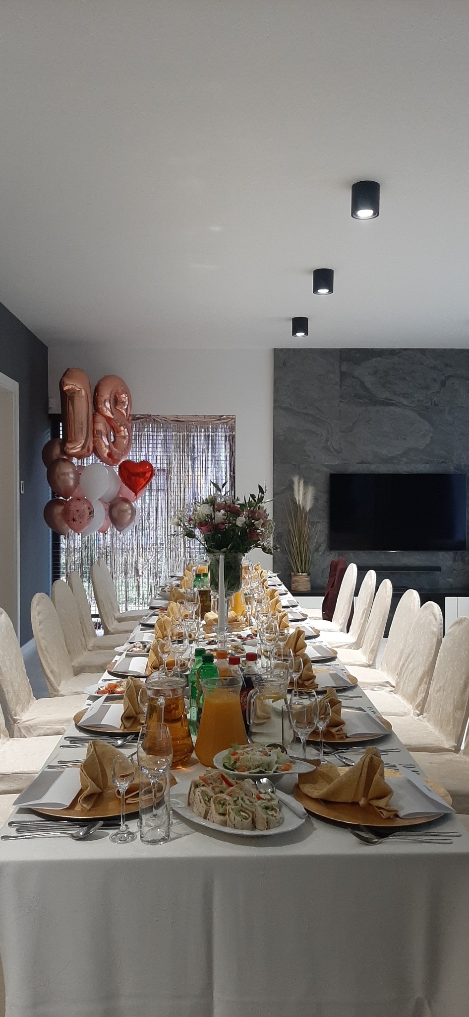 Uroczysty stół na osiemnastkę: biały obrus, złote dekoracje, balony, kwiaty i napoje. Przygotowany do świętowania w nowoczesnym wnętrzu.