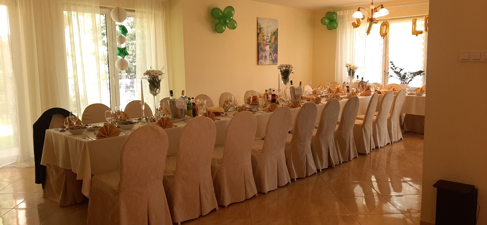 Uroczysta aranżacja stołu z nakryciami, zastawą i dekoracjami na przyjęcie. W tle okno z balonami i napisem 'STO LAT'.