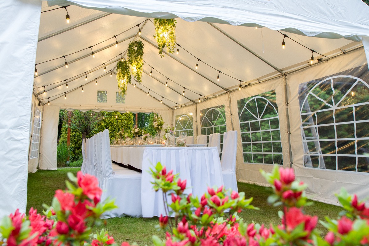 Uroczysty biały namiot z oknami, oświetlony girlandami żarówek, z dekoracjami kwiatowymi i stołem nakrytym do przyjęcia, widziany zza kwitnących krzewów.