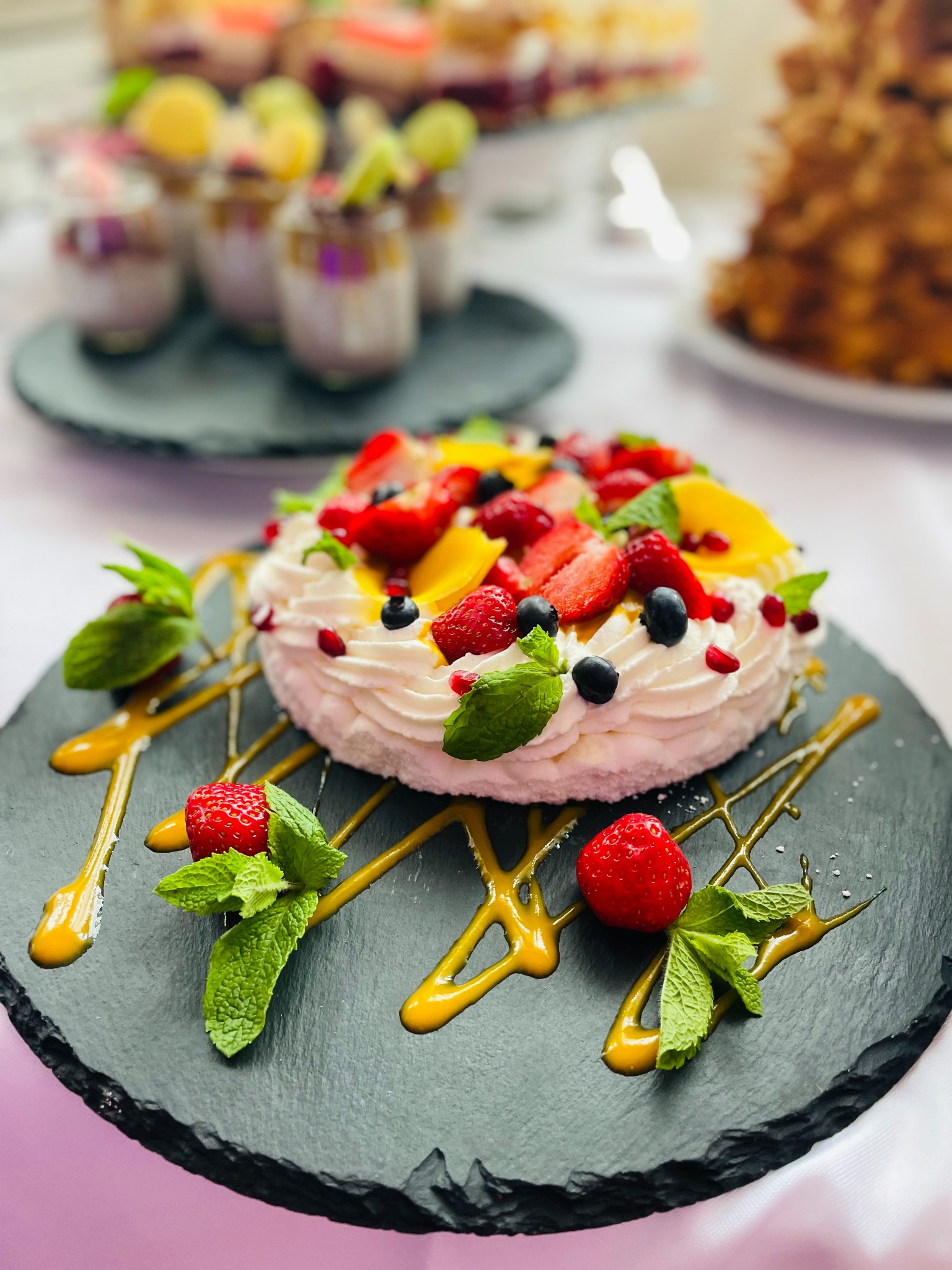 Elegancko udekorowany tort Pavlova z truskawkami, malinami i mango na czarnym talerzu, z dodatkiem sosu i mięty, w tle desery w szklankach i tort.