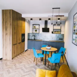 Nowoczesna kuchnia z drewnianą zabudową, szarymi szafkami, okrągłym stołem z turkusowymi krzesłami i parkietem ułożonym w jodełkę, oświetlona wiszącymi lampami.