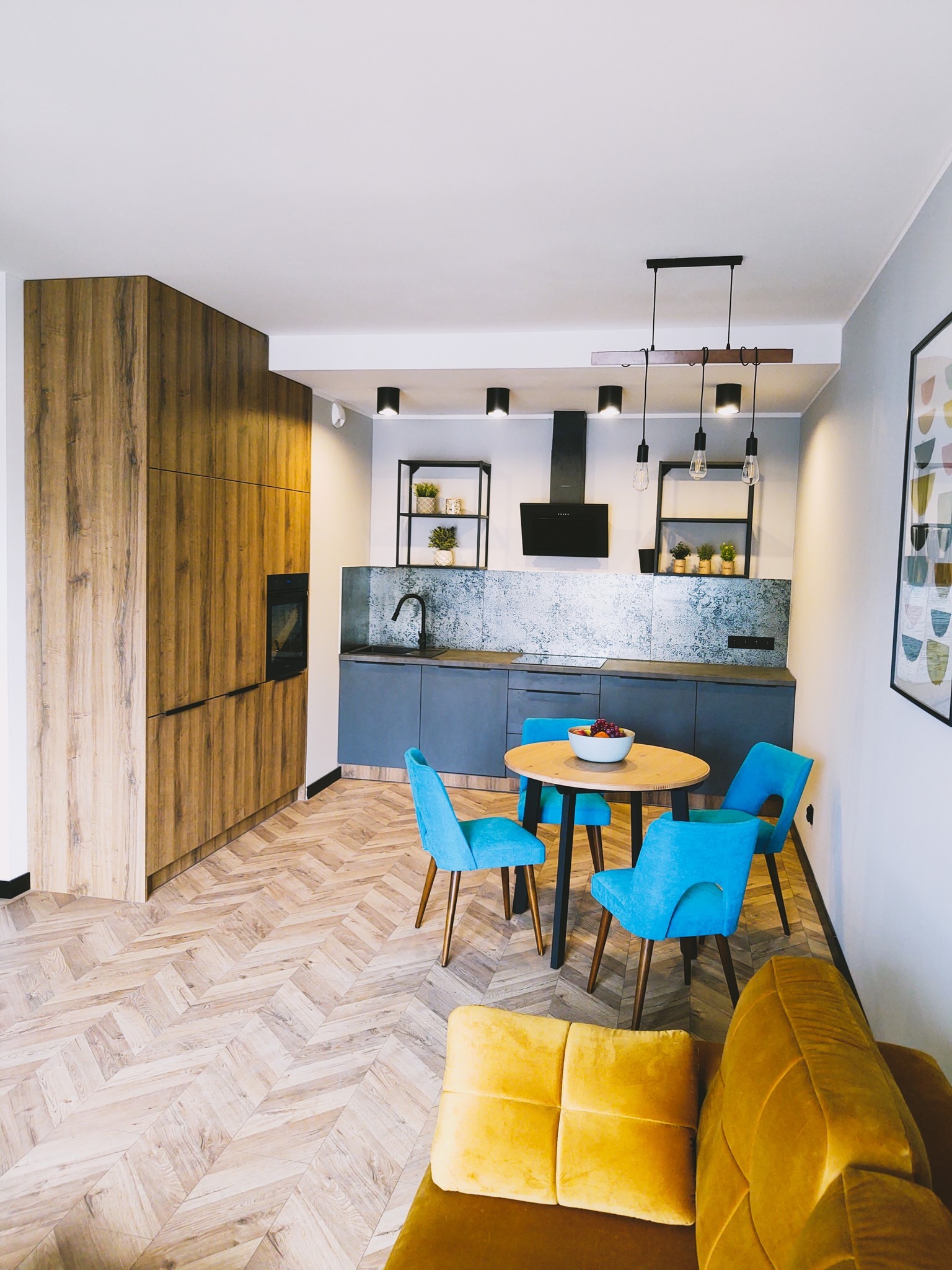 Nowoczesna kuchnia z drewnianą zabudową, szarymi szafkami, okrągłym stołem z turkusowymi krzesłami i parkietem ułożonym w jodełkę, oświetlona wiszącymi lampami.