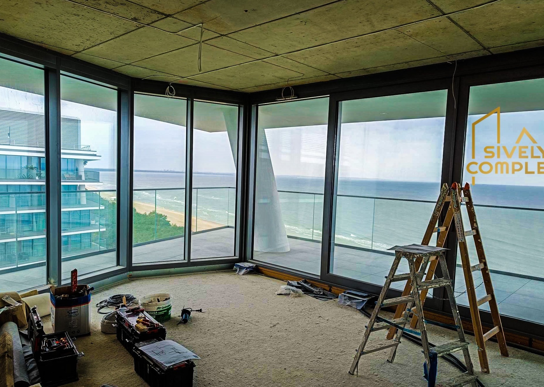 Wnętrze apartamentu z widokiem na morze w trakcie prac wykończeniowych, widoczne narzędzia, drabiny i surowy betonowy sufit z wystającymi kablami.