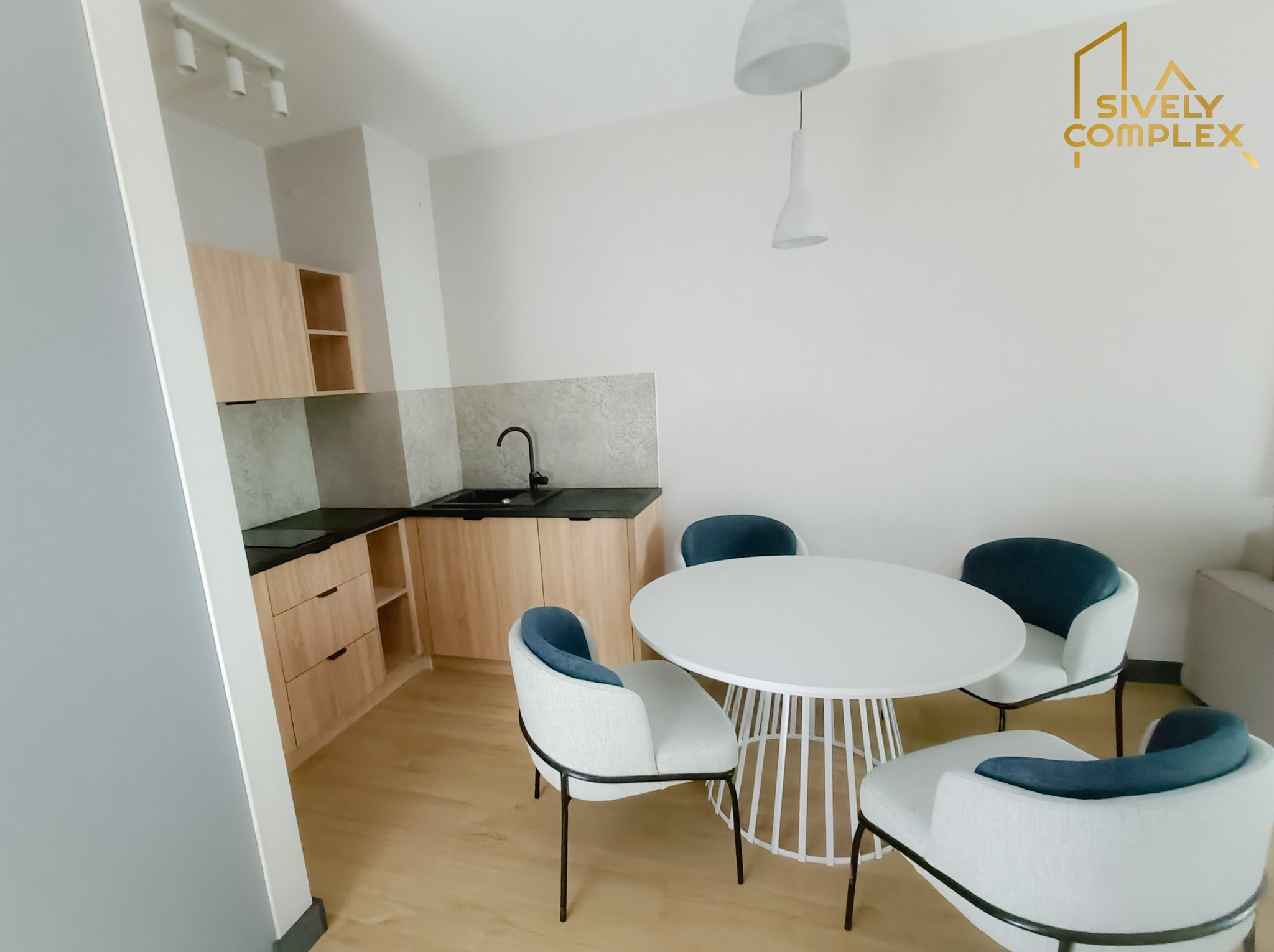 Jasna kuchnia z drewnianymi szafkami, czarnym blatem i okrągłym białym stołem z krzesłami z niebieskimi akcentami, oświetlona lampą wiszącą i reflektorami.