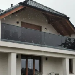 Nowoczesny balkon z szklaną balustradą w grafitowym odcieniu, widok z dołu podczas opadów śniegu, na tle białej elewacji domu z ciemną dachówką.