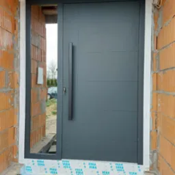 Nowoczesne, grafitowe drzwi wejściowe z długim, pionowym uchwytem, zamontowane w surowym stanie budynku z widoczną czerwoną cegłą i folią ochronną na ramie.