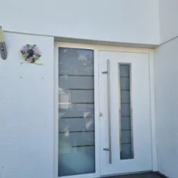 Białe drzwi wejściowe z długim, pionowym uchwytem i przeszklonym panelem, ozdobione kwiatowym wiankiem przy ścianie budynku z teksturą.