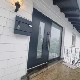 Nowoczesne, grafitowe drzwi wejściowe z przeszkleniami, zamontowane w białej elewacji z drewnianą okładziną, skrzynka na listy obok.