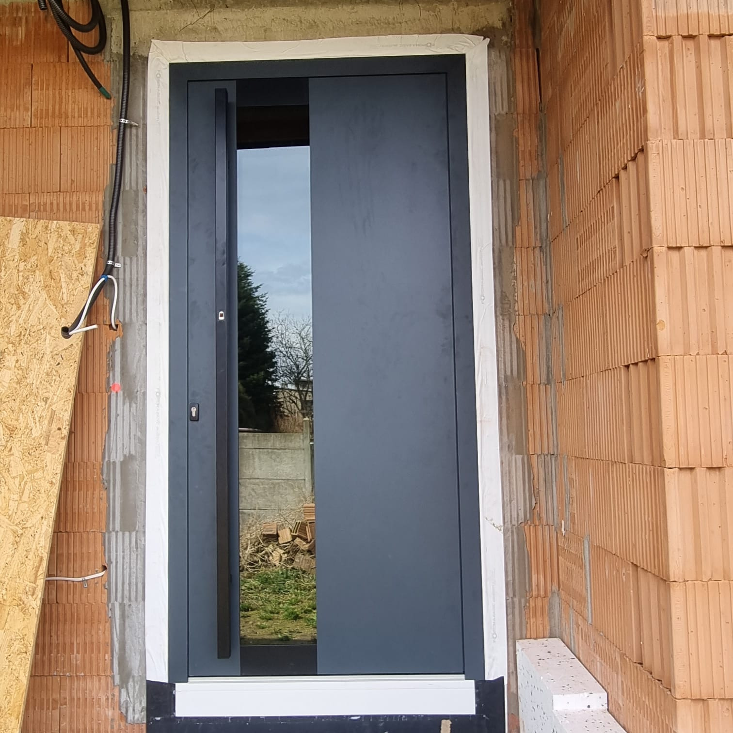 Montaż nowoczesnych, grafitowych drzwi wejściowych z przeszkleniem, widoczne surowe wykończenie muru z cegły ceramicznej i instalacje elektryczne.