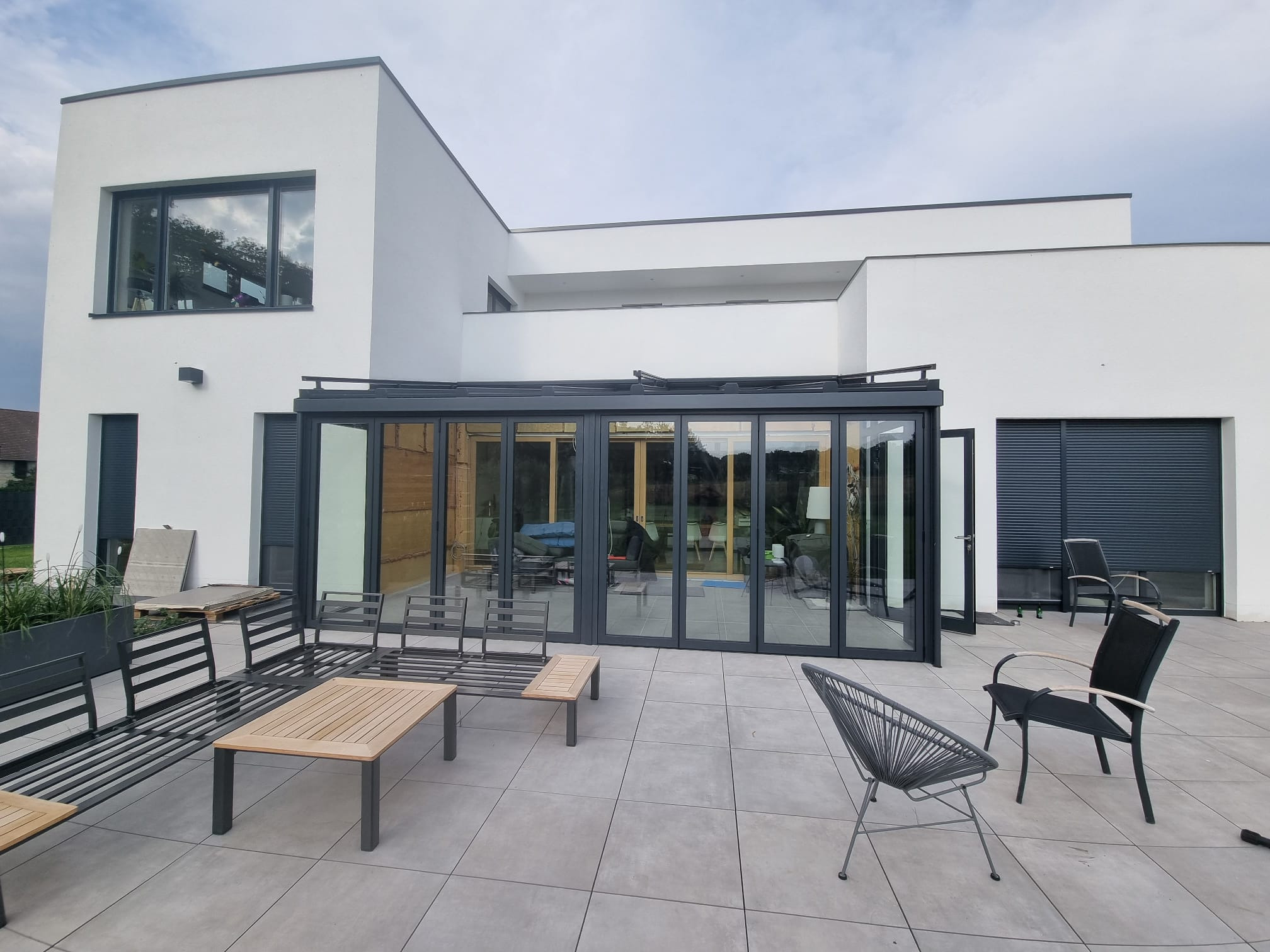 Nowoczesny dom z dużym tarasem, na którym znajdują się meble ogrodowe, widoczne przesuwne szklane drzwi tarasowe w ciemnej ramie oraz okno z roletami zewnętrznymi.