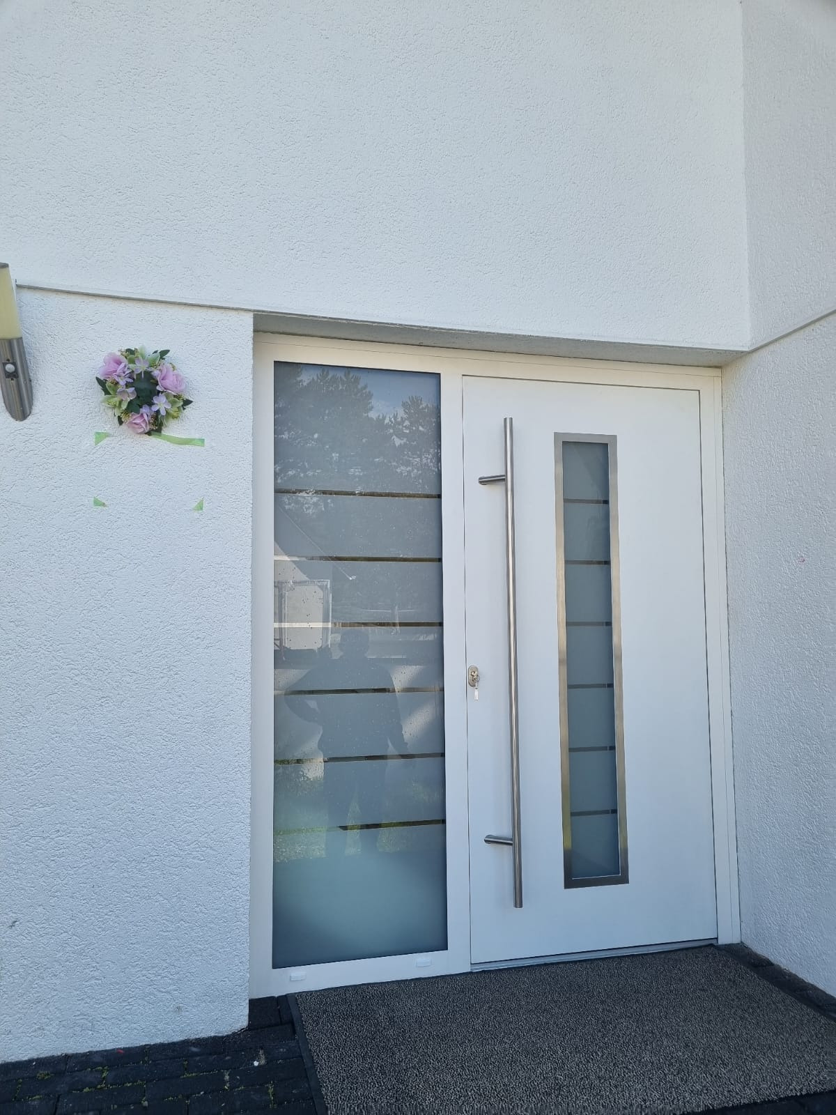 Białe drzwi wejściowe z długim, pionowym uchwytem i przeszklonym panelem, ozdobione kwiatowym wiankiem przy ścianie budynku z teksturą.