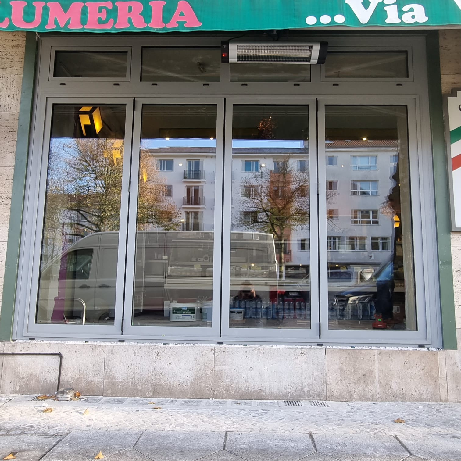 Szare, czteroskrzydłowe okno wystawowe z widocznym odbiciem ulicy i budynku naprzeciwko, nad oknem grzejnik elektryczny, na chodniku leżą liście.