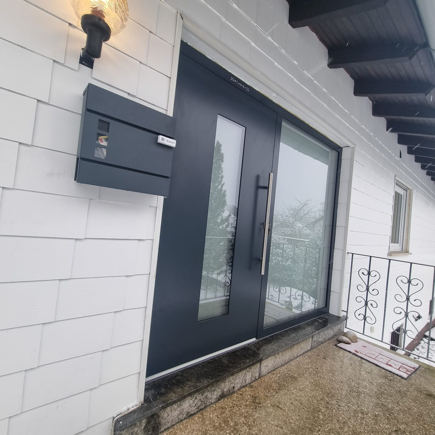 Nowoczesne, grafitowe drzwi wejściowe z przeszkleniami, zamontowane w białej elewacji z drewnianą okładziną, skrzynka na listy obok.