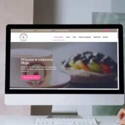 Ekran komputera wyświetla stronę internetową cukierni z Jodłownika, prezentującą ofertę tortów i ciast. Dłoń użytkownika nawiguje po stronie za pomocą myszki.