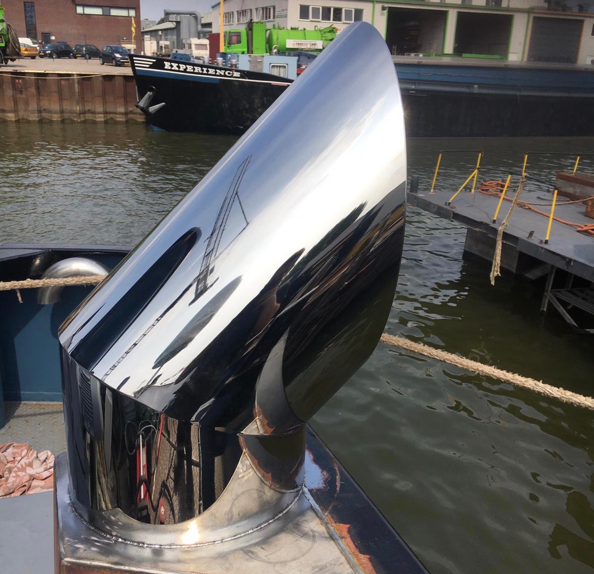 Błyszczący, nierdzewny komin na łodzi, odbijający otoczenie portowe i fragment drabiny, z liną cumowniczą w dolnej części kadru.