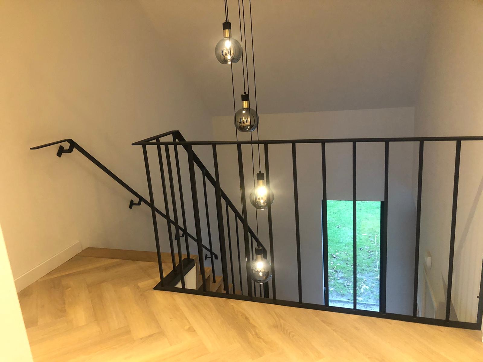 Nowoczesna, czarna balustrada schodowa z metalowymi słupkami i poręczą, oświetlona wiszącymi lampami z żarówkami Edisona, widok z góry na parkiet ułożony w jodełkę.