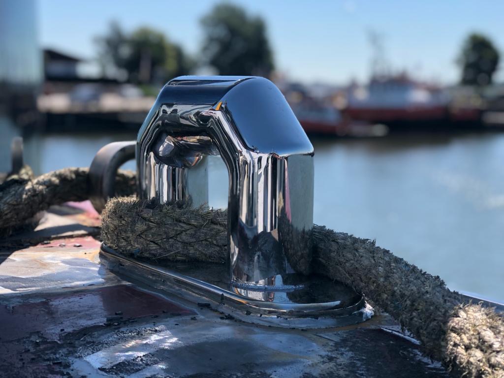 Błyszcząca, chromowana knaga cumownicza na pokładzie łodzi, z grubą liną cumowniczą, na tle wody i innych łodzi.