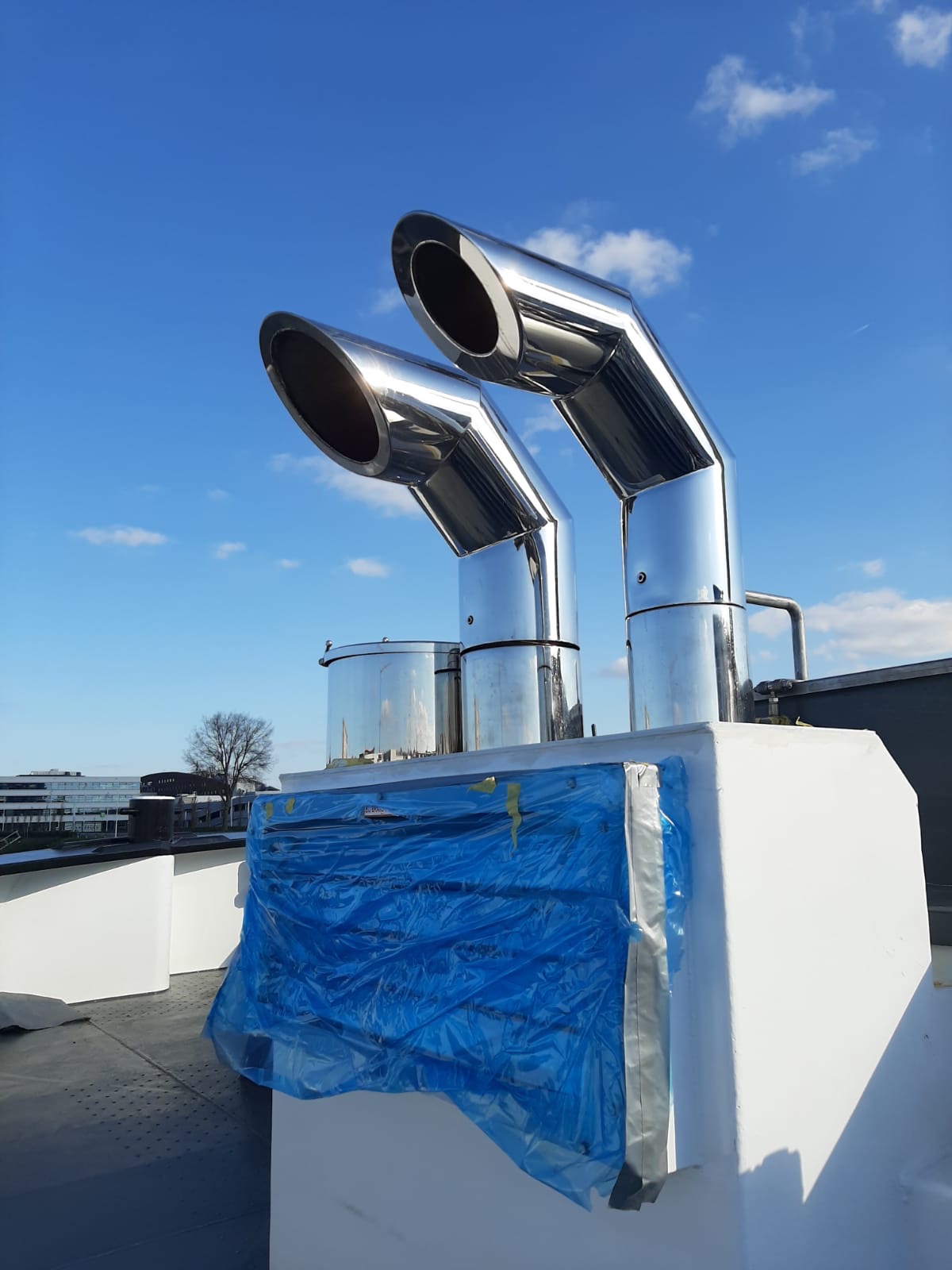 Dwa błyszczące, nierdzewne kominy wentylacyjne na dachu, zabezpieczone niebieską folią ochronną, na tle błękitnego nieba z lekkimi chmurami.