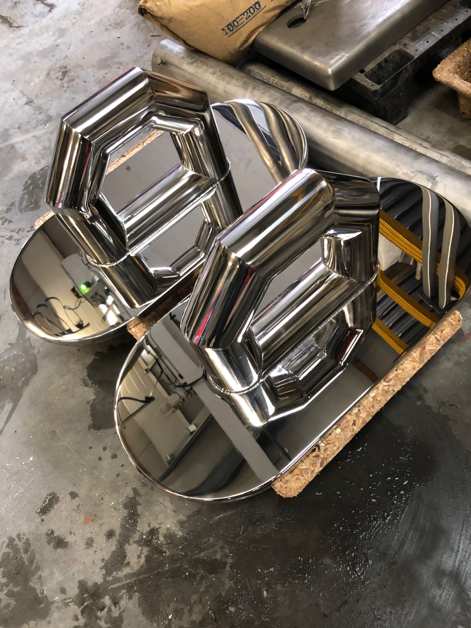 Dwa błyszczące, nierdzewne elementy konstrukcyjne o geometrycznym kształcie, prawdopodobnie fragmenty balustrady lub poręczy, leżące na kawałkach korka na betonowej podłodze warsztatu.