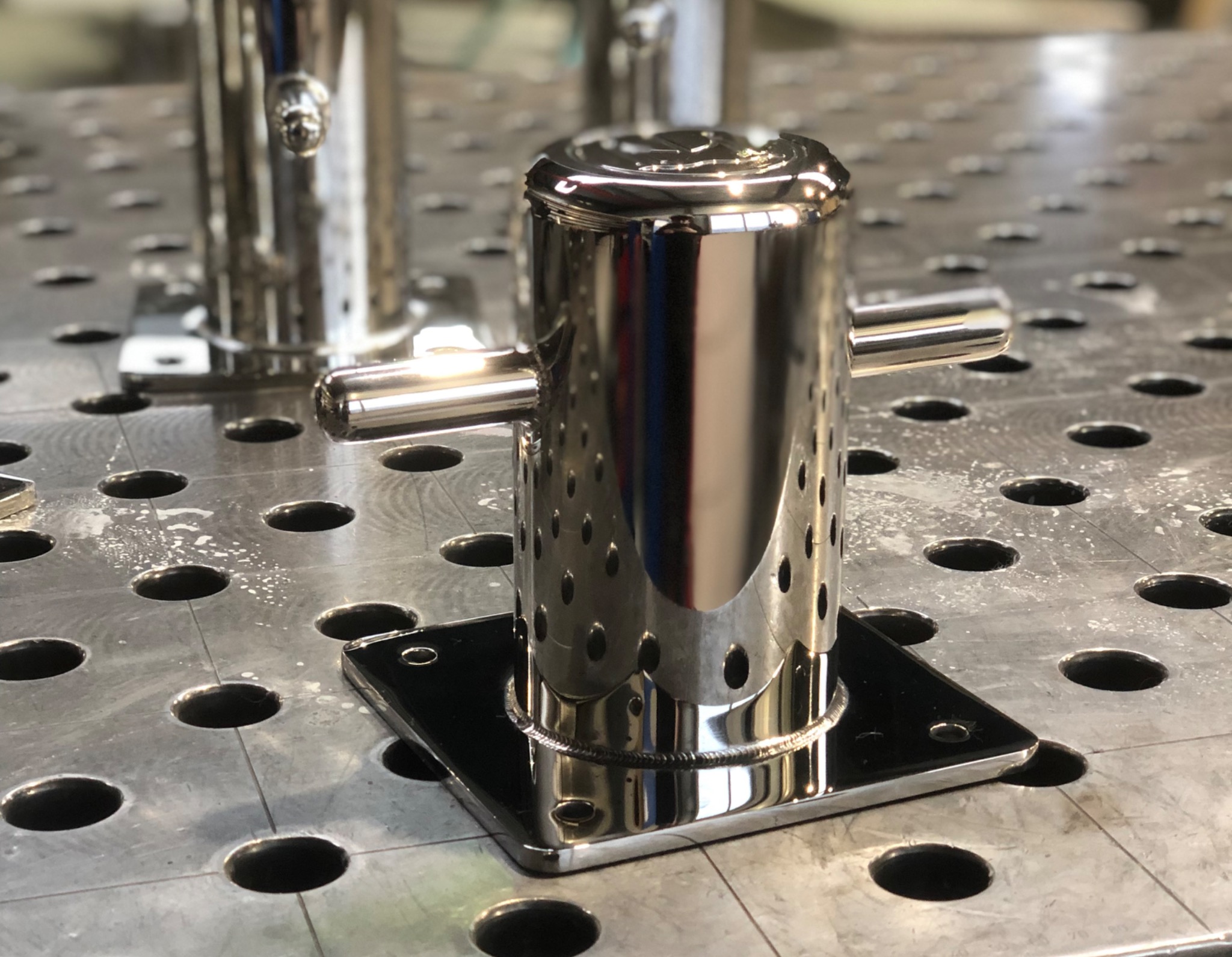 Błyszczący, spawany element ze stali nierdzewnej z otworami, umieszczony na perforowanym stole spawalniczym. Widoczne ślady po spawaniu i odbicia światła.