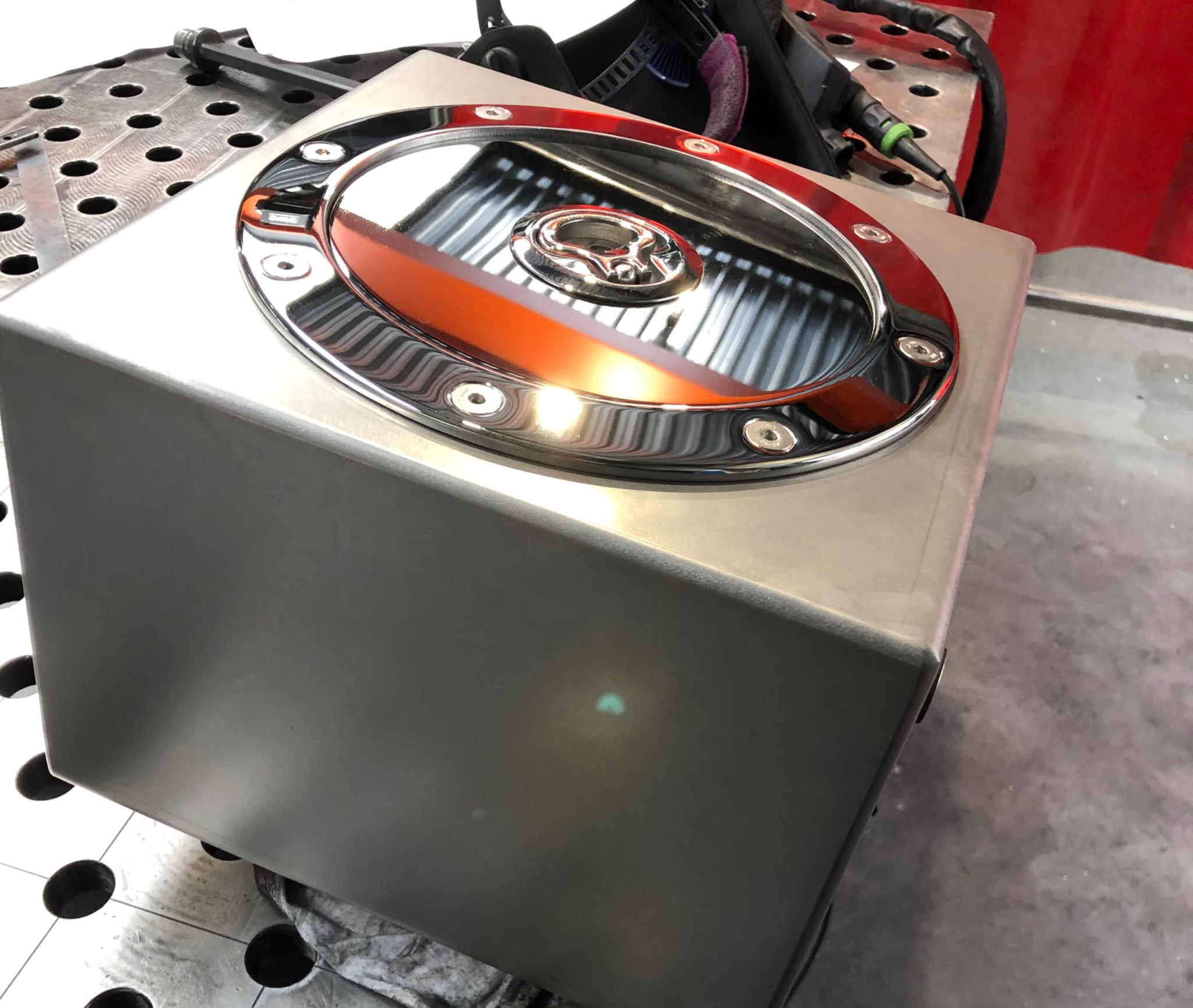 Kwadratowa obudowa ze stali nierdzewnej z okrągłym, chromowanym deklem na wierzchu, przymocowanym śrubami, leżąca na perforowanym stole spawalniczym.