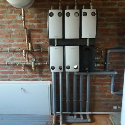 Goman Energy Solutions - Sześć nowoczesnych grzejników panelowych zamontowanych na ceglanej ścianie, z widocznymi rurami doprowadzającymi wodę oraz szafką z kluczykiem poniżej.