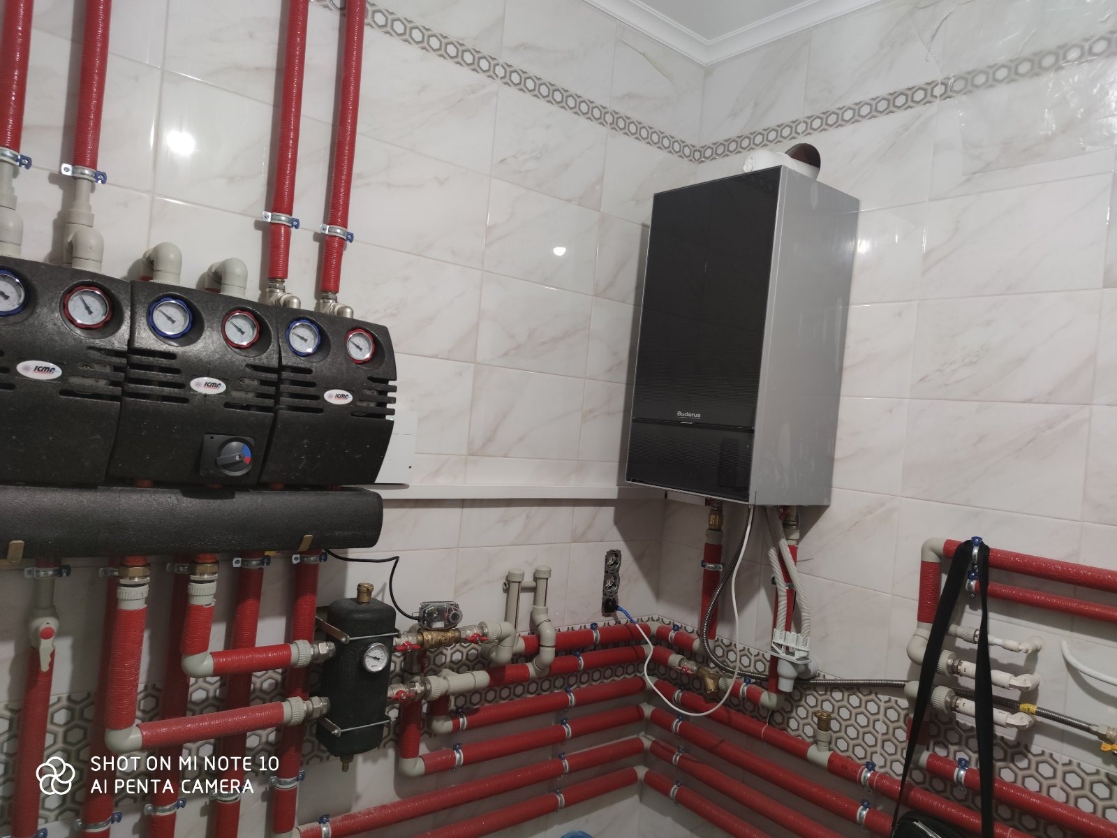 Kocioł grzewczy Buderus zamontowany na ścianie w pomieszczeniu z rozdzielaczem i rurami w czerwonej izolacji, widoczne manometry i zawory.