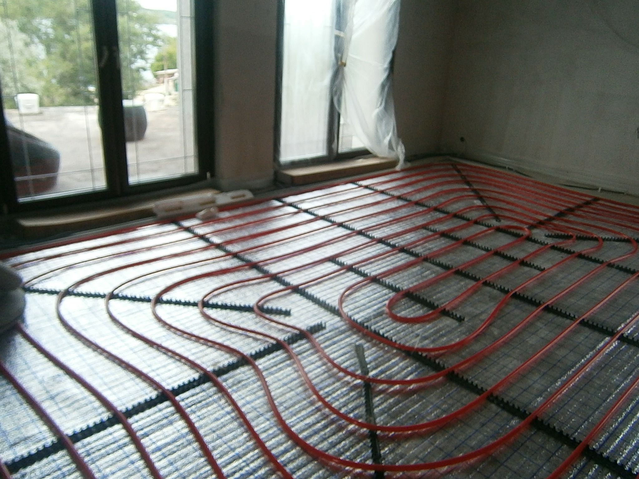 Montaż ogrzewania podłogowego: czerwone rurki ułożone na srebrnej folii izolacyjnej w pomieszczeniu z oknami.