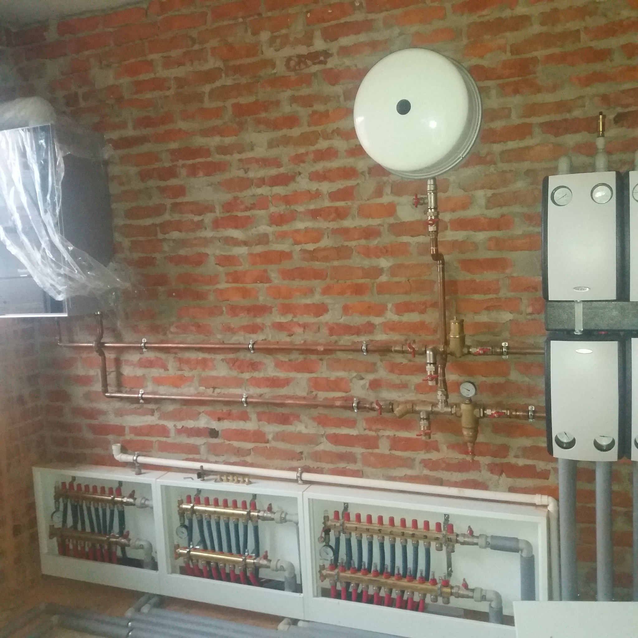 Rozdzielacze ogrzewania podłogowego z rurami miedzianymi i kotłami gazowymi na tle ściany z czerwonej cegły.