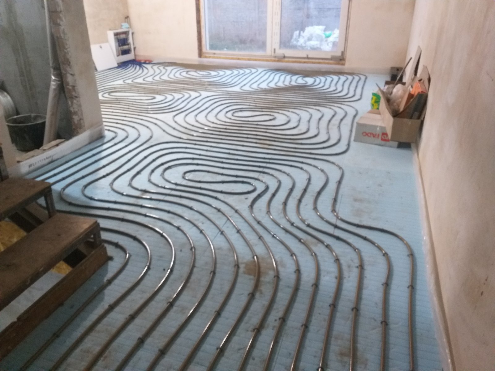 Rury systemu grzewczego ułożone spiralnie na izolacji podłogowej w trakcie instalacji w surowym pomieszczeniu, widoczne narzędzia i karton.