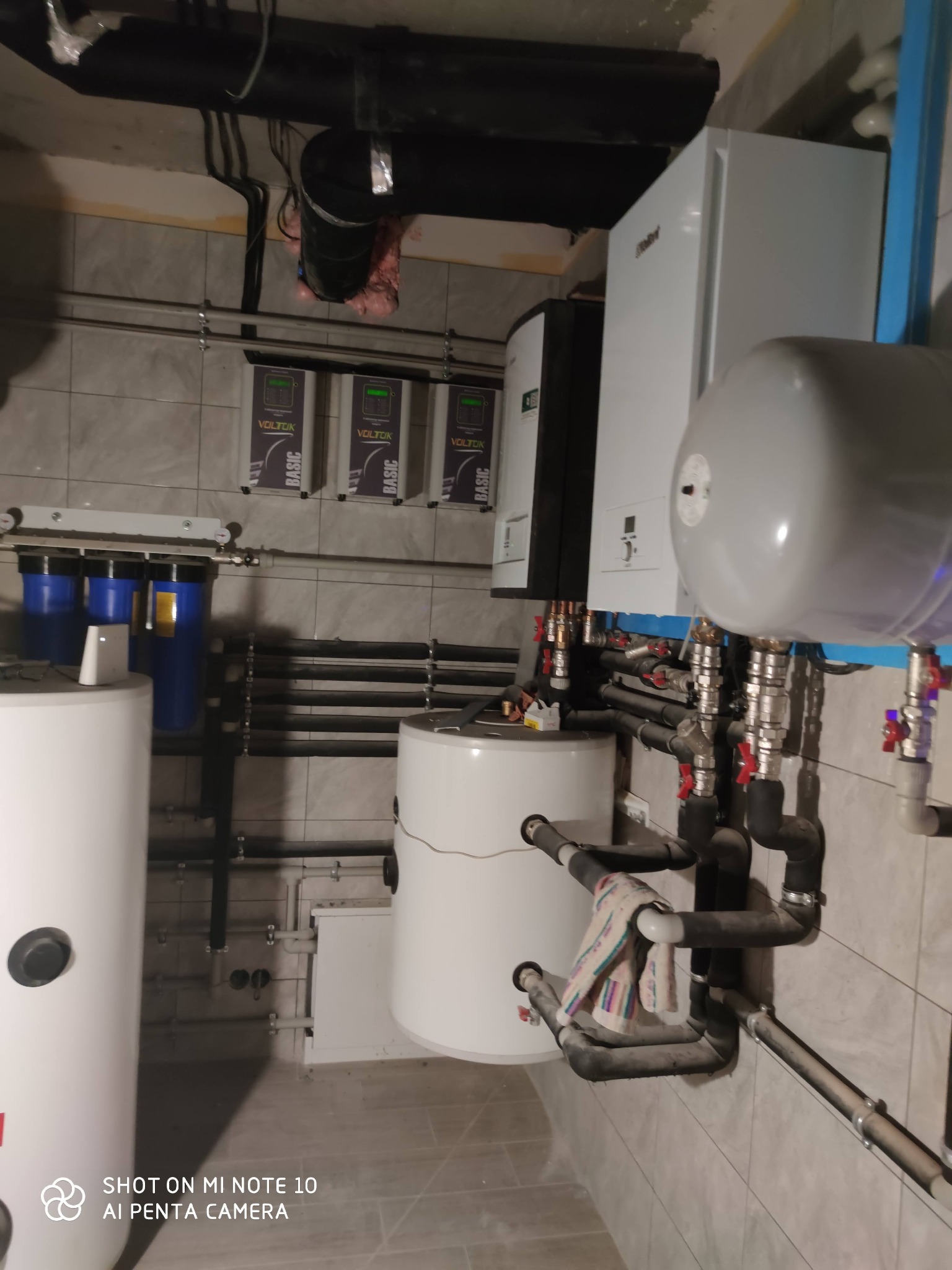 Kotłownia z białymi zbiornikami, rurami, filtrami wody i sterownikami w obudowach VOLTOIK BASIC na ścianie z szarymi płytkami.