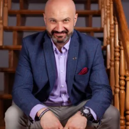 Uśmiechnięty mężczyzna z brodą, ubrany w niebieską marynarkę w kratę i liliową koszulę, siedzi na drewnianych schodach, prezentując zegarek i bransoletkę z napisem.