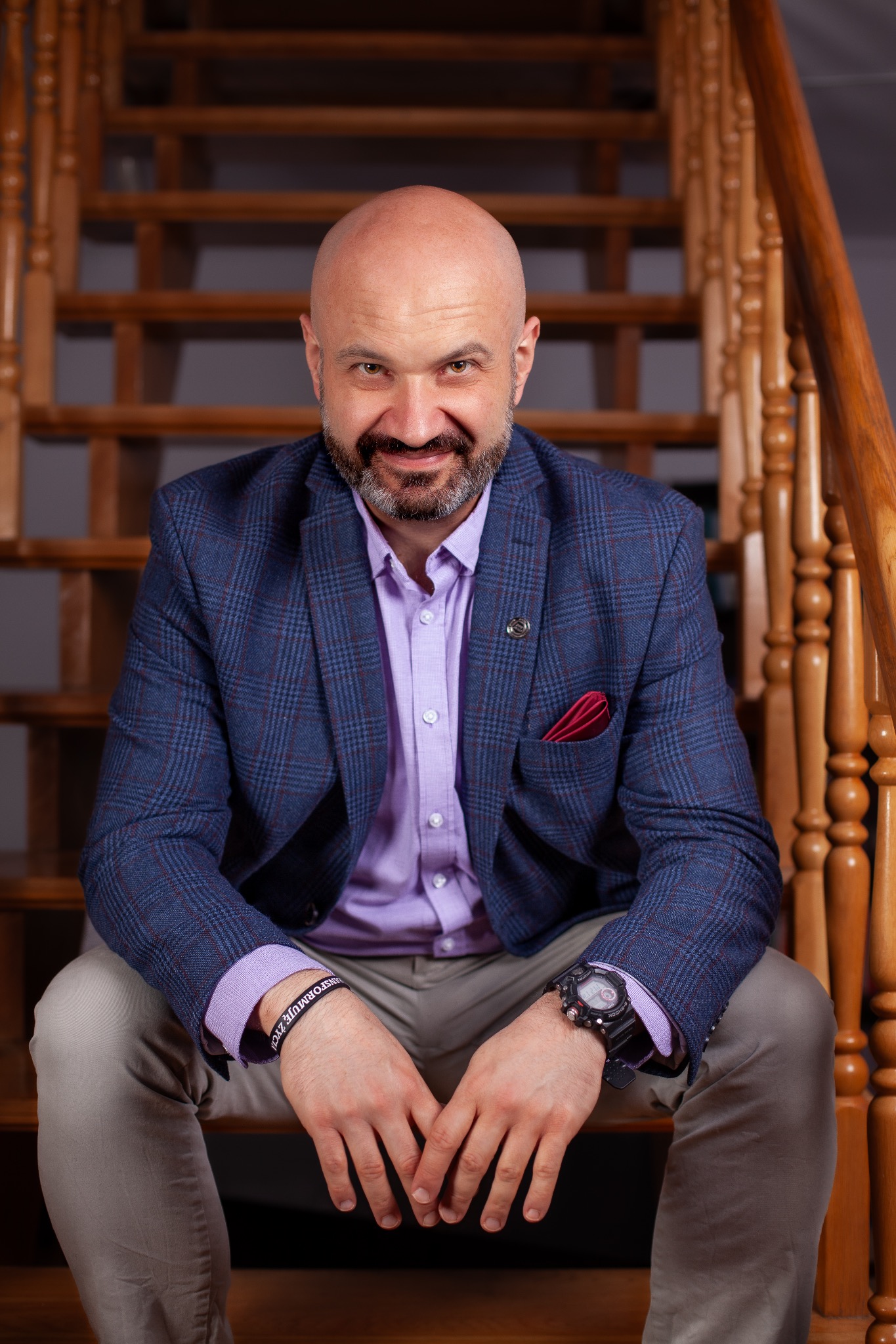 Uśmiechnięty mężczyzna z brodą, ubrany w niebieską marynarkę w kratę i liliową koszulę, siedzi na drewnianych schodach, prezentując zegarek i bransoletkę z napisem.