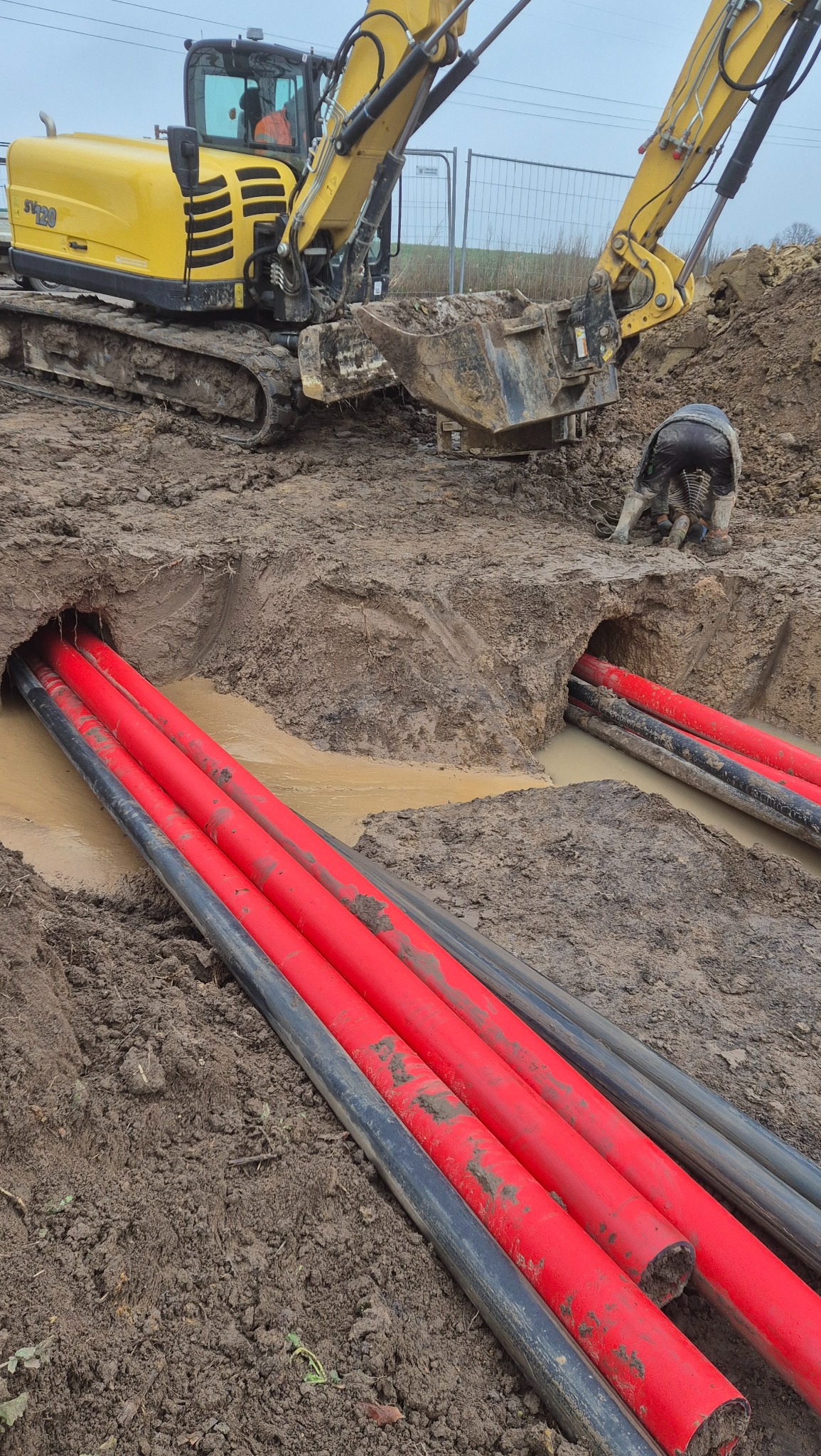 Żółta koparka i pracownik przy układaniu czerwonych i czarnych rur w wykopie wypełnionym wodą. Widoczne błoto i ziemia.