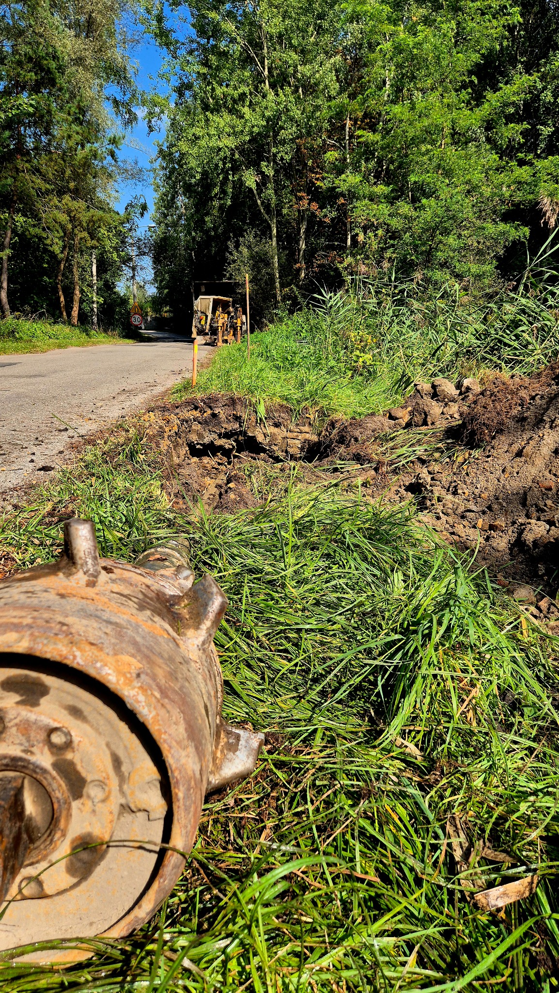 Wykonany wykop pod drogą z widoczną maszyną do przewiertów sterowanych i wykopaną ziemią. W tle droga i zieleń.