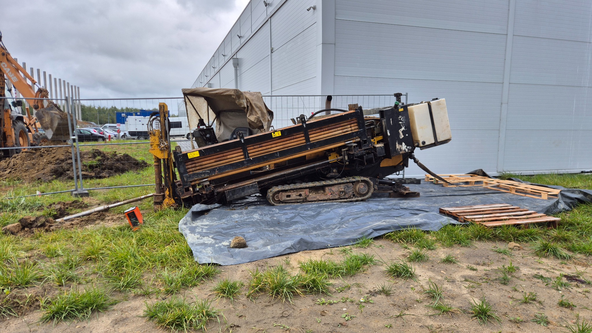 Maszyna do przewiertów sterowanych na placu budowy, widoczna koparka i hala w tle. Sprzęt ustawiony na folii i paletach, przygotowany do pracy.