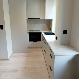 RB Studio Projekty Rafał Bartkowski - Jasna kuchnia na wymiar z drewnianą podłogą. Ułożenie w kształcie litery L, czarne uchwyty szafek, płyta grzewcza i zlew. Minimalistyczny design.