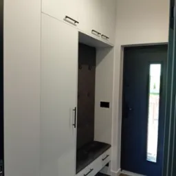 Biała szafa wnękowa z siedziskiem i wieszakami, czarne uchwyty, minimalistyczny design, widok na drzwi wejściowe. Nowoczesne rozwiązanie do przedpokoju.