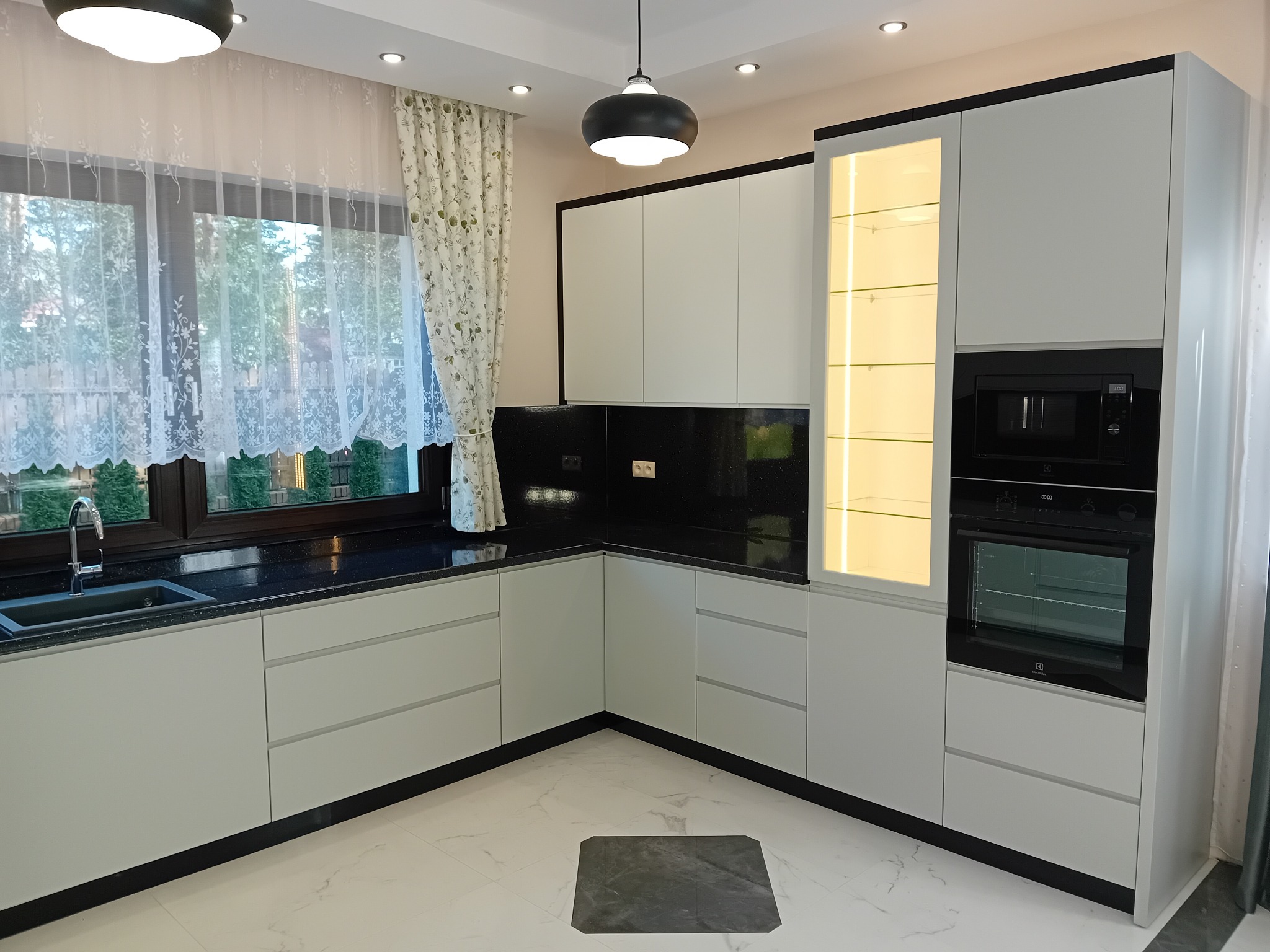 Nowoczesna kuchnia z białymi szafkami, czarnym blatem i oświetleniem LED, uzupełniona o czarne akcenty na obrzeżach i armaturze, z widokiem na ogród przez okno z delikatnymi firankami.