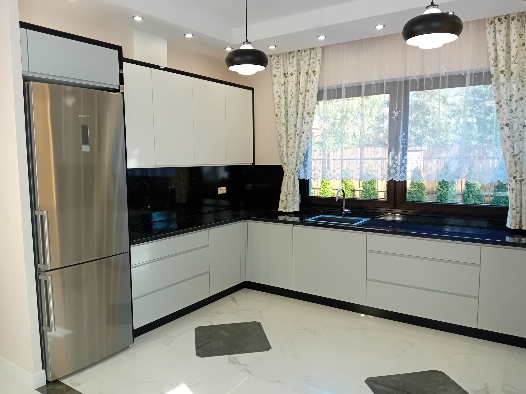 Nowoczesna kuchnia z białymi szafkami, czarnym blatem z połyskiem i marmurową podłogą z ciemnymi wstawkami, oświetlona wiszącymi lampami i światłem dziennym przez okno z delikatnymi firankami.